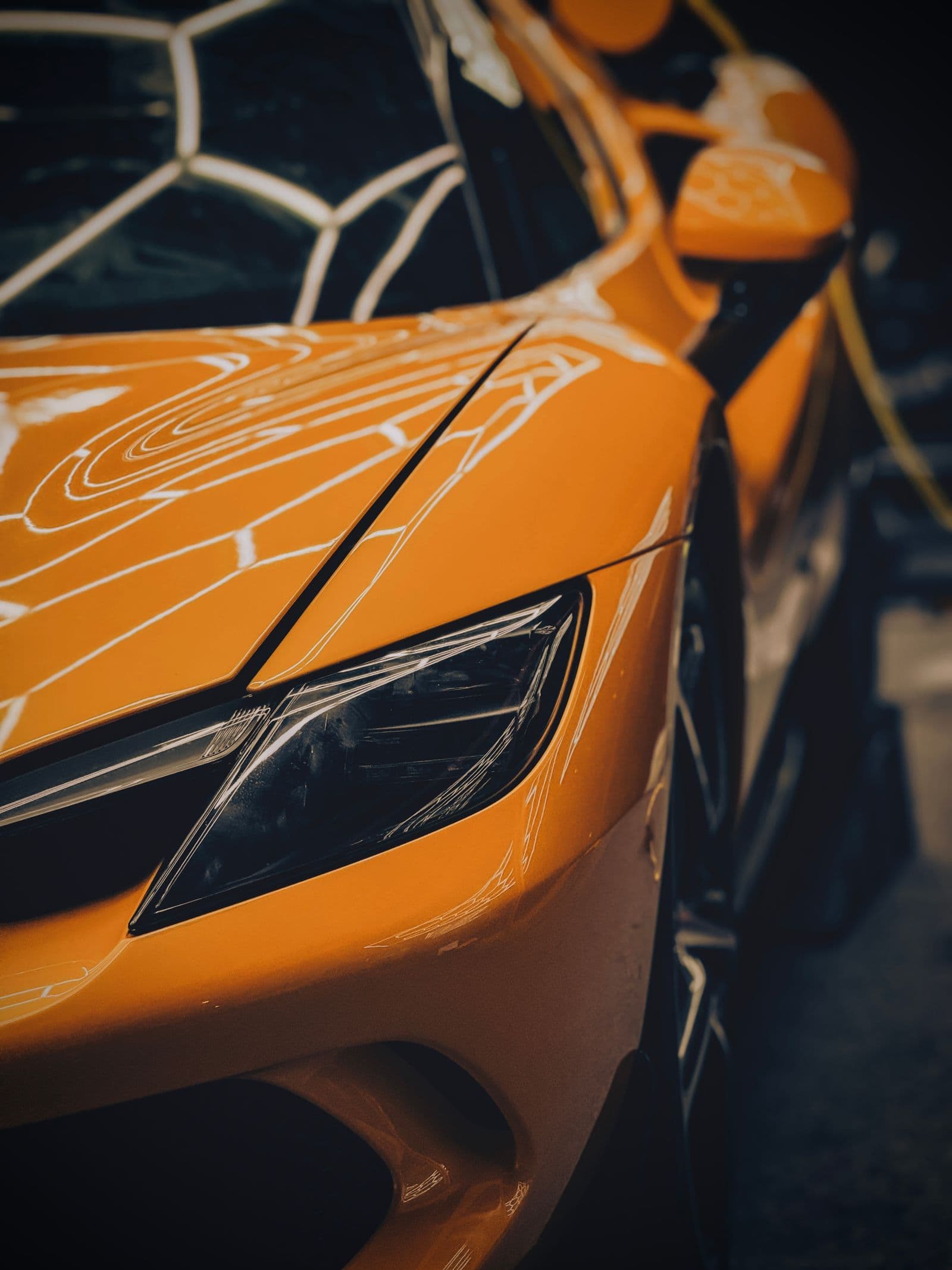 A glossy orange sports car front, its hood mirroring an overhead structure